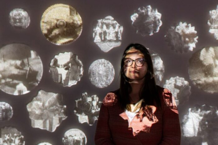 An image of Katina with images in light projected on her face and body and the dark screen she is standing in front of.