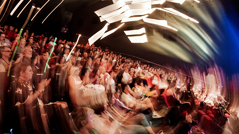 A view of an audience in a theater, the image distorted by a lens into rippling circles.