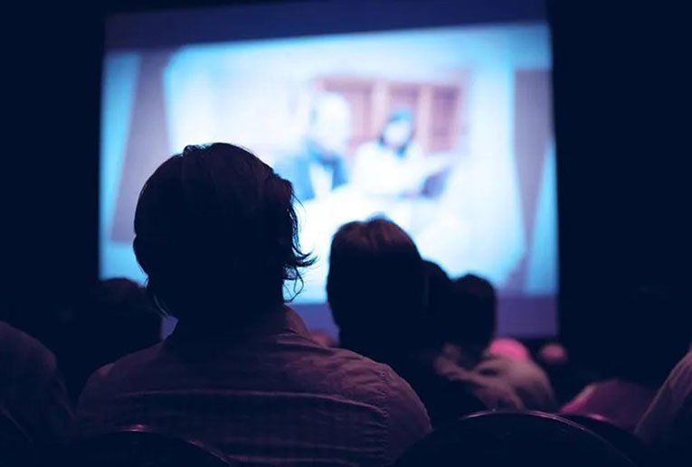 Interior of a darkened theater shot from behind the audience members, who are in silhouette, looking at the screen, which has a blurry image of two people on it.