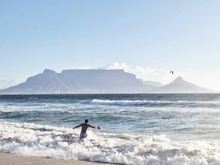 A sunny beach's surf, and a man running into the water. In the background are the distinctive mountains of Cape Town.