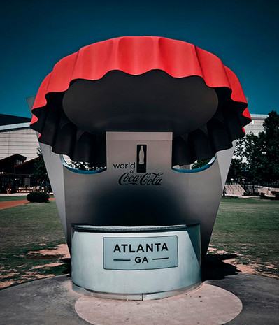 A statue of a large Coca-Cola bottle cap. On the bottle cap opener holding the cap it says World of Coca-Cola, and below that on the pedestal it say Atlanta GA.