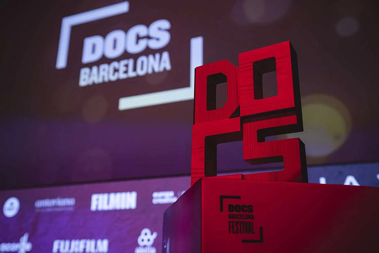 The logo for Docs Barcelona Festival is in the right foreground, carved DOCS stacked like blocks on top of a red box that also has the logo in black on it. In the background, the theater screen has DOCS Barcelona framed in a half box.