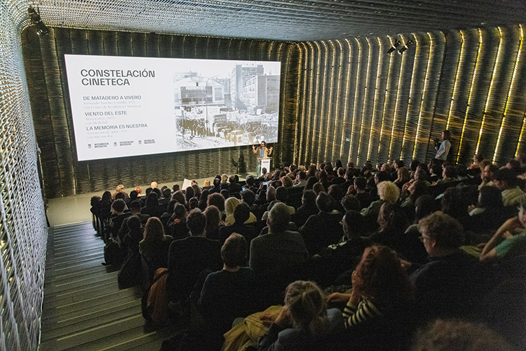 An audience in a darkened theater looking at a speaker at a podium. Behind the speaker on the screen is a slide that says "Constellation Cineteca."