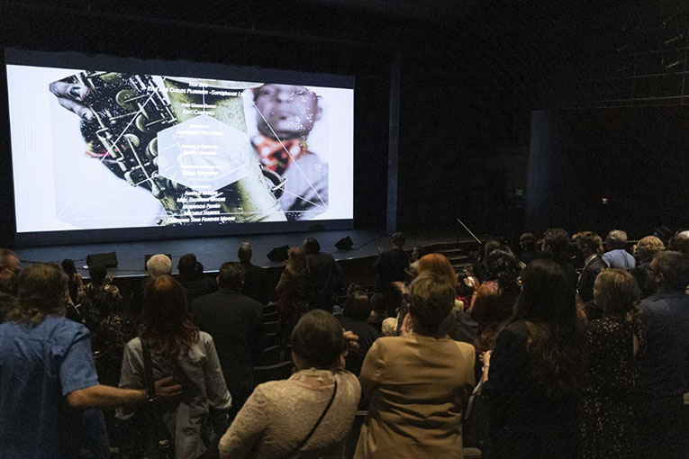 A darkened theater interior shot from behind the audience, who are standing and looking at an image on the screen. The image is a man's face on the right and a close-up of part of saxophone's keys on the left.