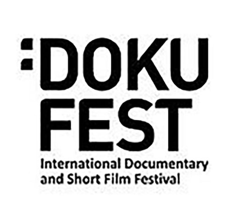 The logo of the festival in black letters on a white background. The first line of text is a colon, then the word DOKU in all caps, then FEST in all caps below it. Below that in smaller font are the words International Documentary and Short Film Festival.