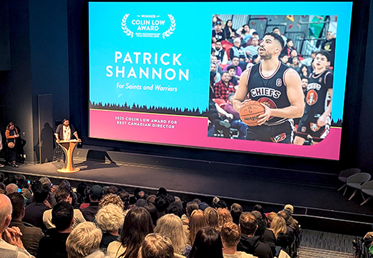 Award ceremony at DOXA film festival, with an audience in a darkened theater. On the screen is a still of a film about basketall player Patrick Shannon that won an award.