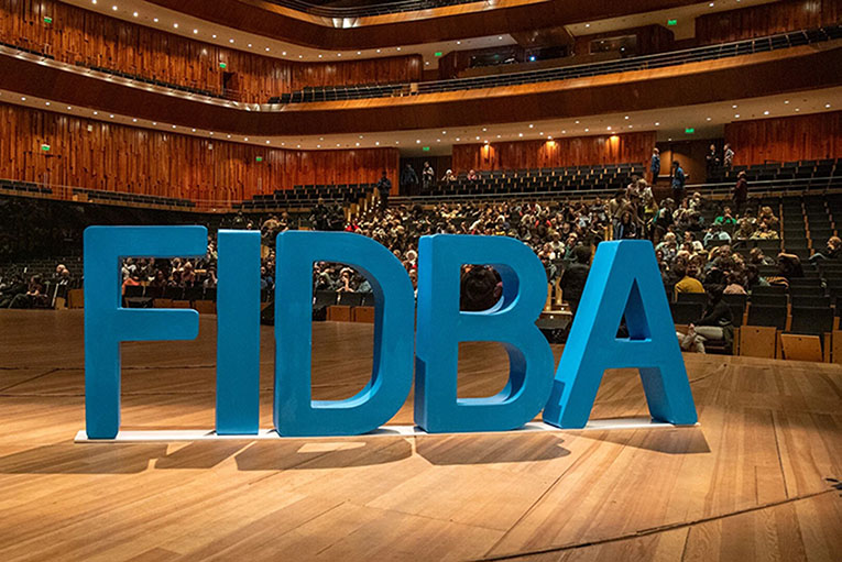 FIDBA logo in large blue letters standing on a stage in an auditorium, an audience in the seats behind it.