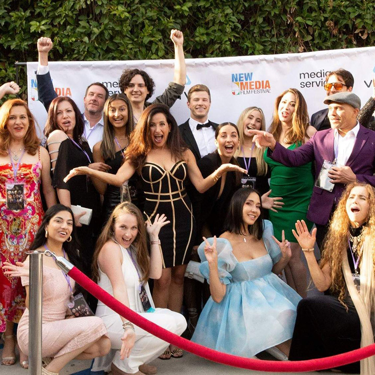 Exterior daytime. A group of people smiling for the camera in front of a white screen with the New Media Film Festival logo on it.