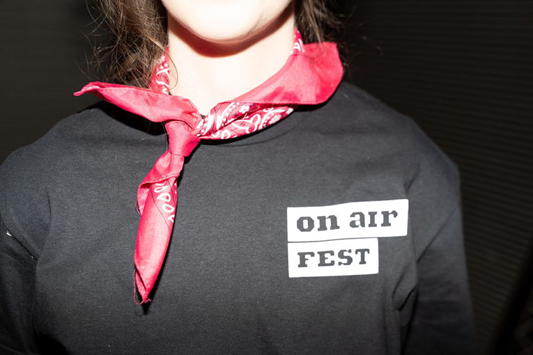 Close-up shot of a woman wearing a red bandanna and an On Air Fest shirt with the logo on the left side of her chest.