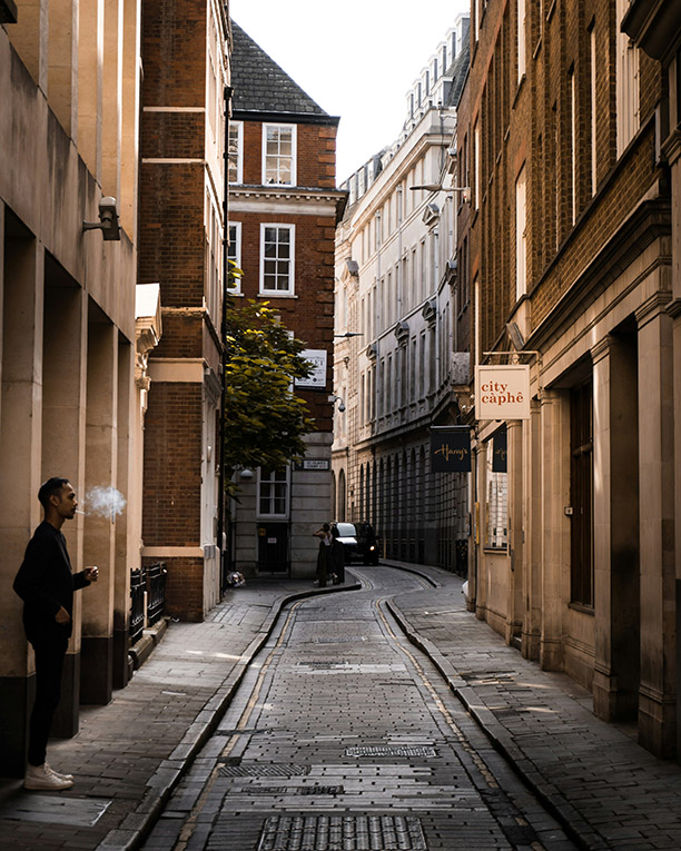 A London street view, a narrow, curving road flanked by tall brick buildings. A man stands against one wall smoking, and a black car is parked at the far end of the road.