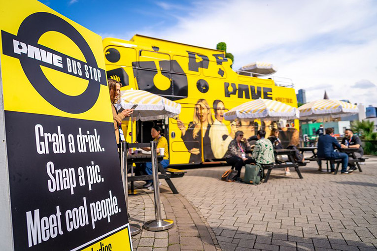 An exterior patio on a sunny day with yellow and black "PAVE" bus in the background. People sit at small tables with yellow and white umbrellas. A yellow, black, and white in the left foreground identifies the space as a PAVE bus stop. Below that the sign says, Grab a drink. Snap a pic. Meet cool people.
