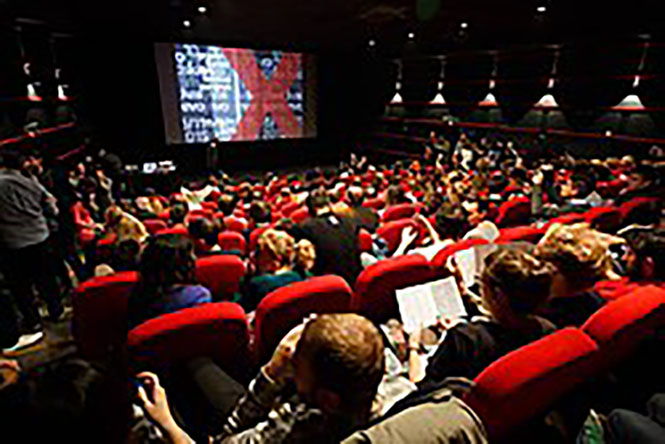 Interior of a theater shot from behind the audience, with a logo on the theater screen.