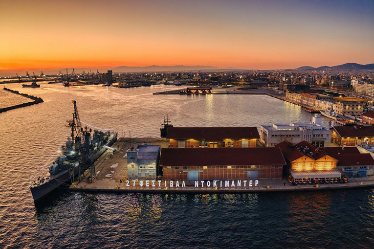 A sunset view of the harbor of Thessaloniki, Greece, with a warship berthed in the foreground alongside a pier with warehouses and other buildings on it, a kind of man-made peninsula. In the background is a view of the city as the city lights are coming on. In the furthest distance there are low mountains.