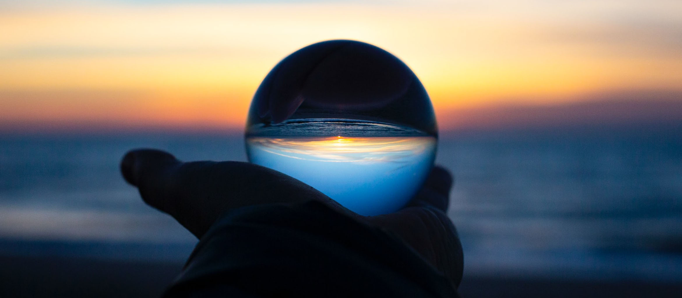 The image shows a hand holding a crystal ball at the beach at sunset. The crystal inverts the image of the sunset and beach, making it look like a galaxy floating in darkness.